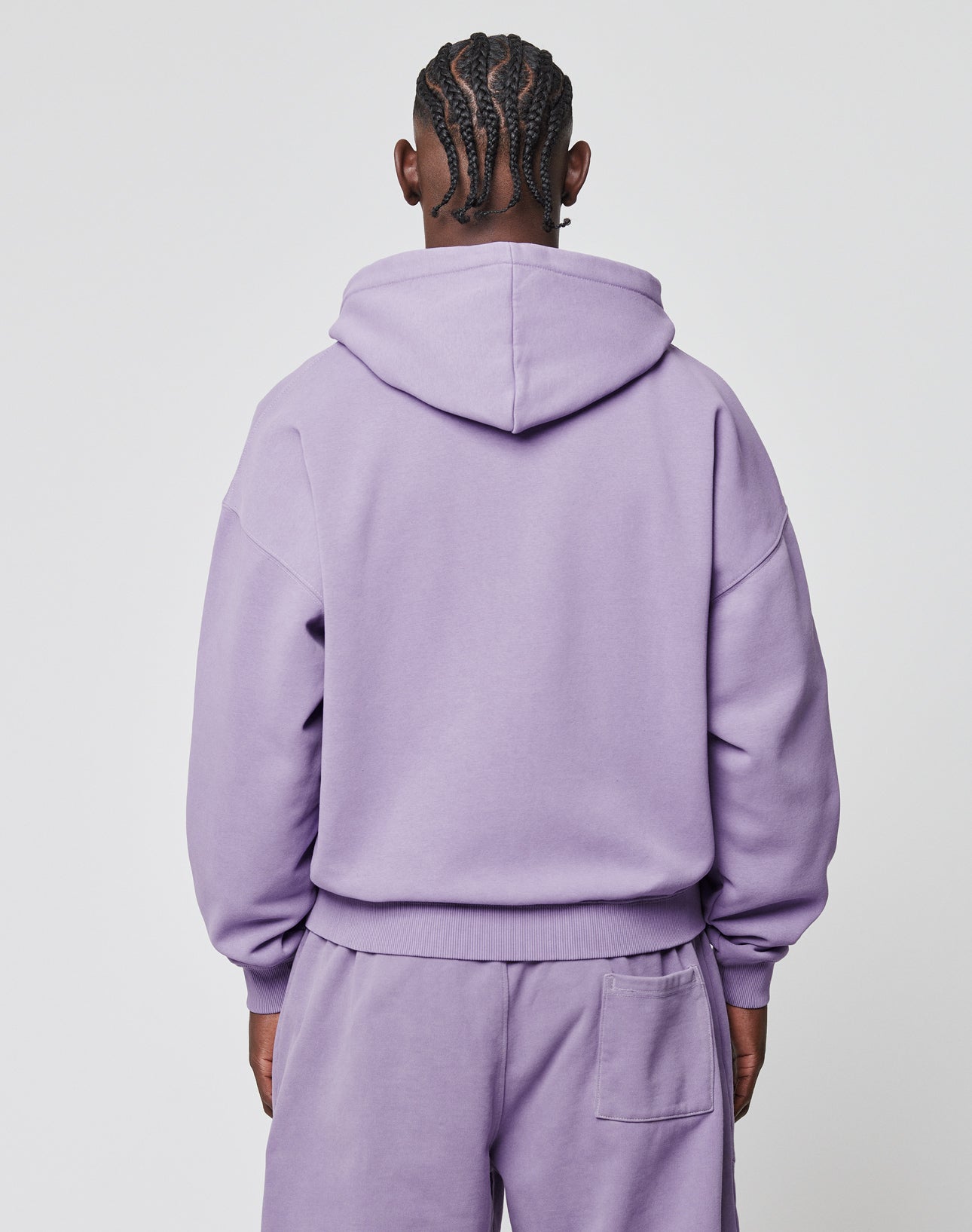 A person with braided hair is seen from behind, wearing the LIVE FAST DIE YOUNG Basic Script Hooded Zip in a matching light purple set with Vintage-Waschung. The background is plain and light-colored.