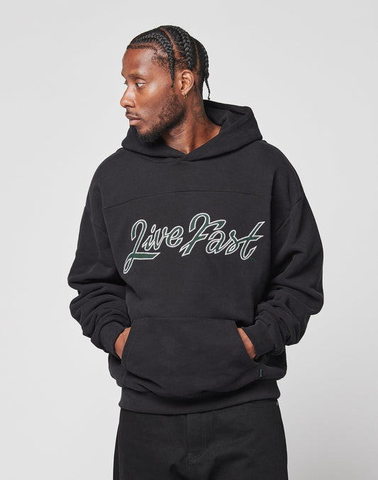 A man with braided hair wears an oversize black LFDY Script Hooded by LIVE FAST DIE YOUNG, “Live Fast” in script on the front, standing hands in pocket and looking left against a plain light background.