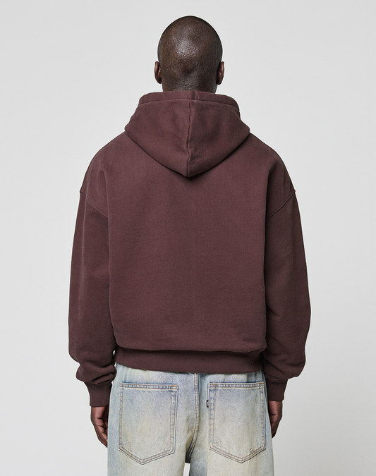 A person with a shaved head is seen from behind, wearing the brown LFDY x Baba Angora Hoodie by LIVE FAST DIE YOUNG and light jeans against a plain gray background—a relaxed, minimalist look inspired by Berliner Stiftung.