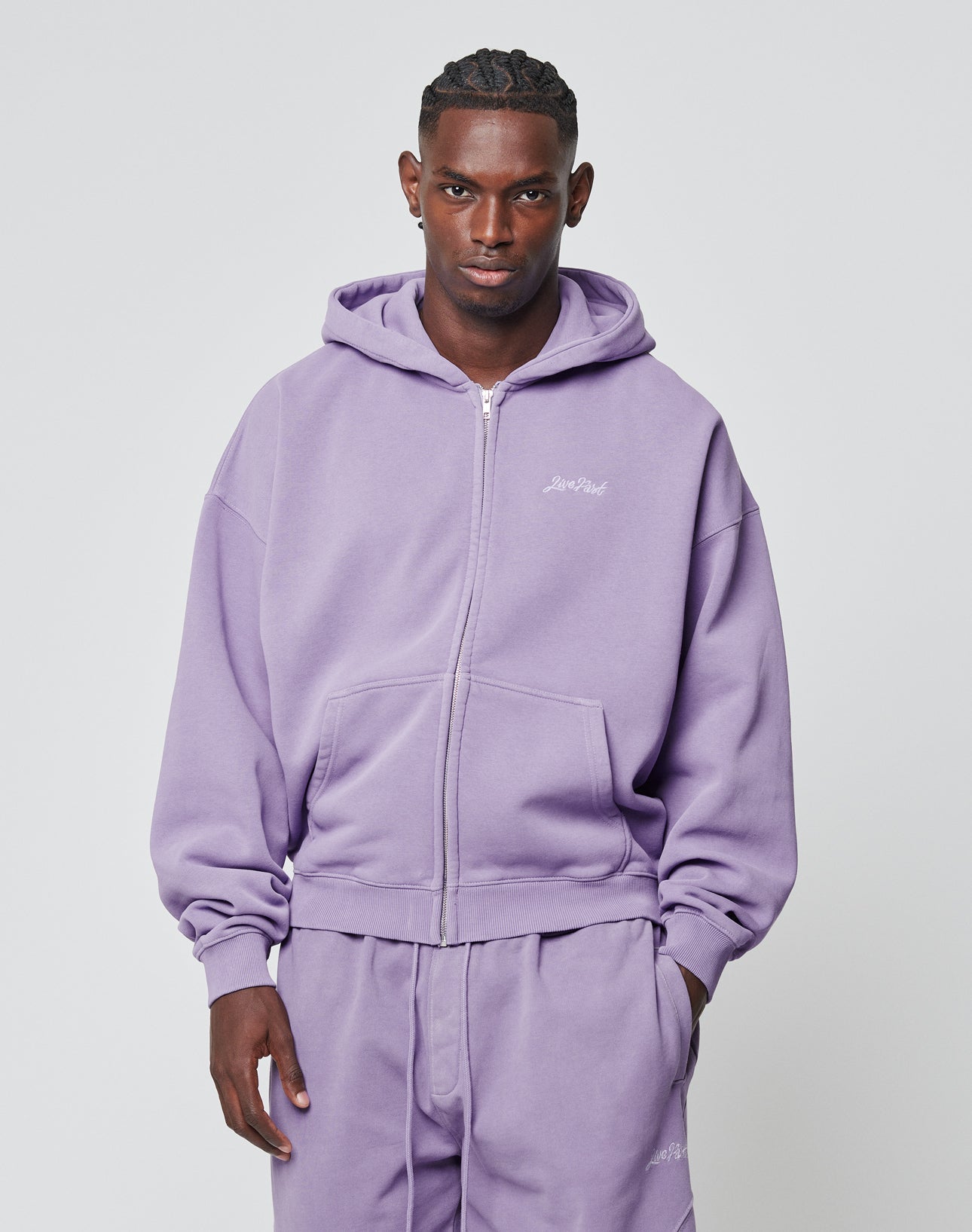 A man faces forward in a matching light purple LIVE FAST DIE YOUNG Basic Script Hooded Zip and sweatpants aus Baumwolle, featuring a small logo embroidery on the left chest. The plain background is light gray.