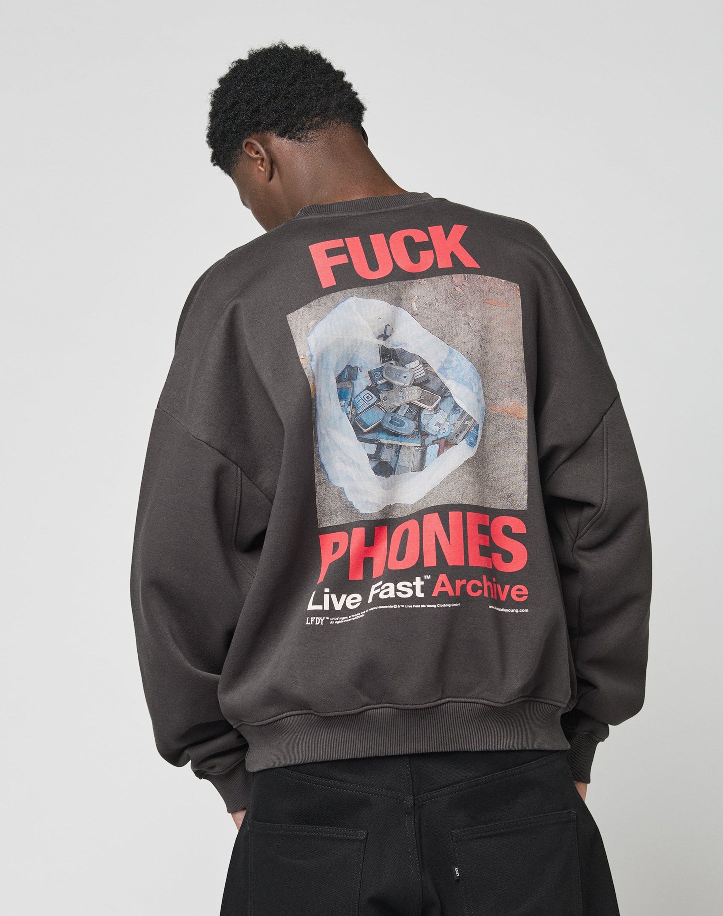 Wearing the LFDY F*ck Sweater by LIVE FAST DIE YOUNG, a person in an oversized black cotton sweatshirt with bold “FUCK PHONES Live Fast Archive” text and discarded phones graphic on the back stands facing away against a plain background.