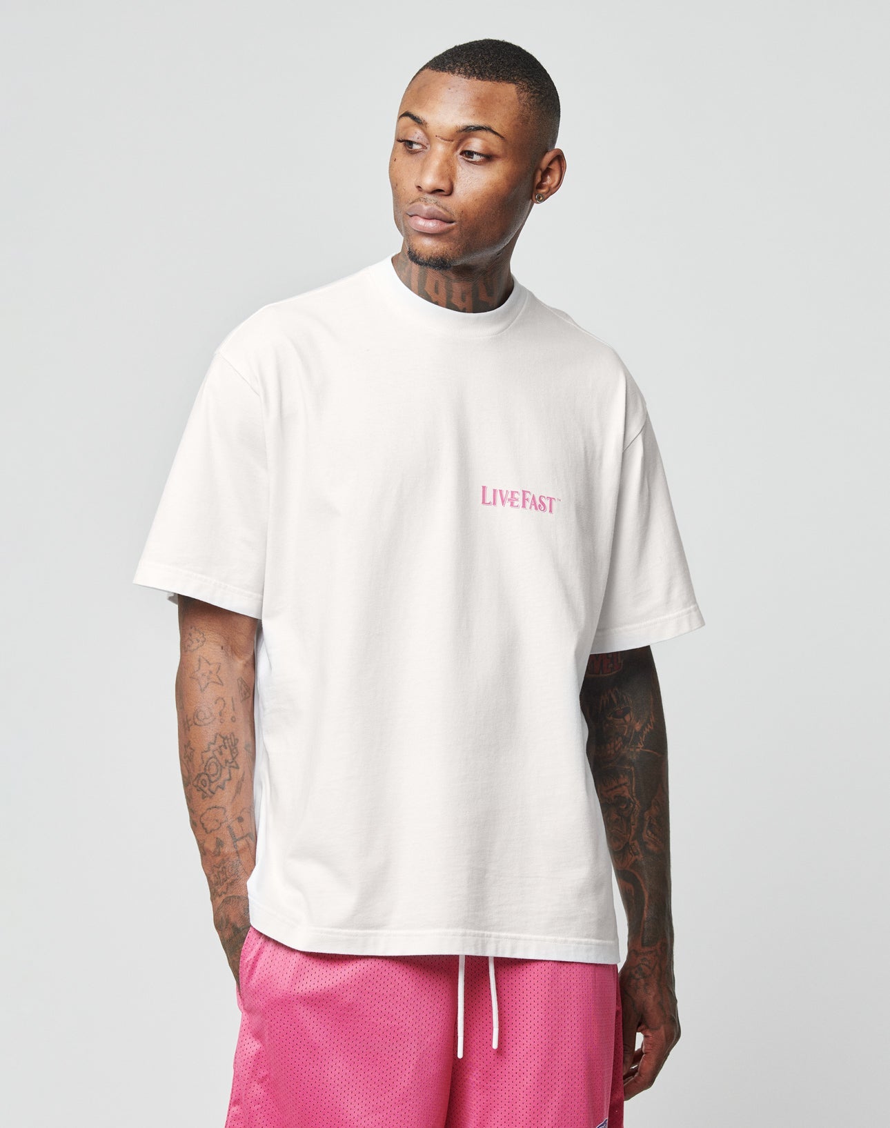 A man wearing a white LFDY Loyal Tee by LIVE FAST DIE YOUNG with an oversize fit and pink shorts stands against a plain light background, looking left with his hands in his pockets.