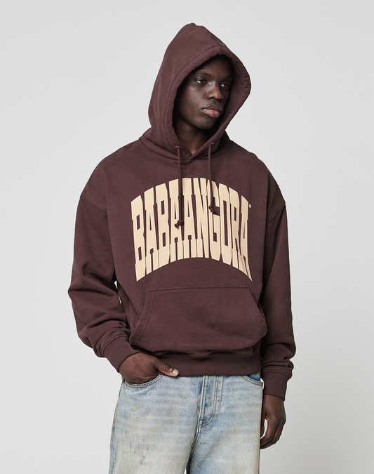 Wearing the LFDY x Baba Angora Hoodie from LIVE FAST DIE YOUNG, with bold beige BABLAGORA print, a person stands against a plain light background in faded blue jeans, one hand in their pocket, looking to the side.