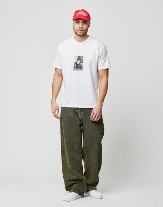 A man in a white graphic tee aus 100% Baumwolle, LFDY Baggy Denim von LIVE FAST DIE YOUNG im dunklen Grünton, weißen Sneakern und roter Baseballcap steht vor weißem Hintergrund und blickt nach vorn.