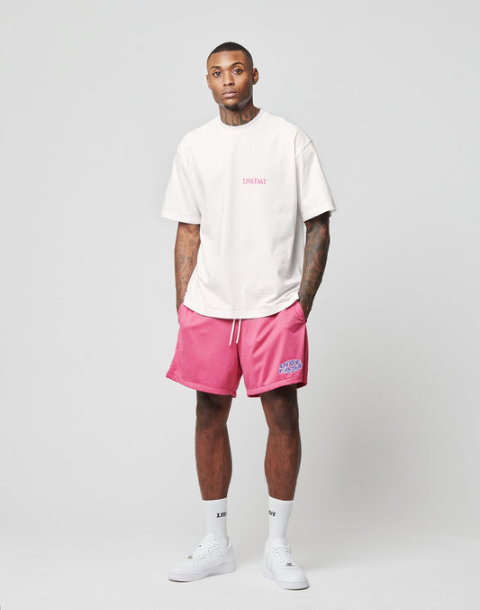 A man stands facing forward in a studio, wearing a white T-shirt and pink LFDY Oval Mesh Shorts by LIVE FAST DIE YOUNG with an elastic waistband. His tattooed arms are relaxed at his sides against a plain light gray background.