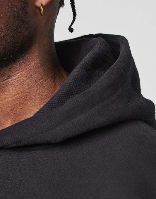 Close-up of a person with brown skin, a small gold hoop earring, and short twists wearing the LIVE FAST DIE YOUNG LFDY Script Hooded black hoodie in Oversize Fit. The background is plain and light-colored.
