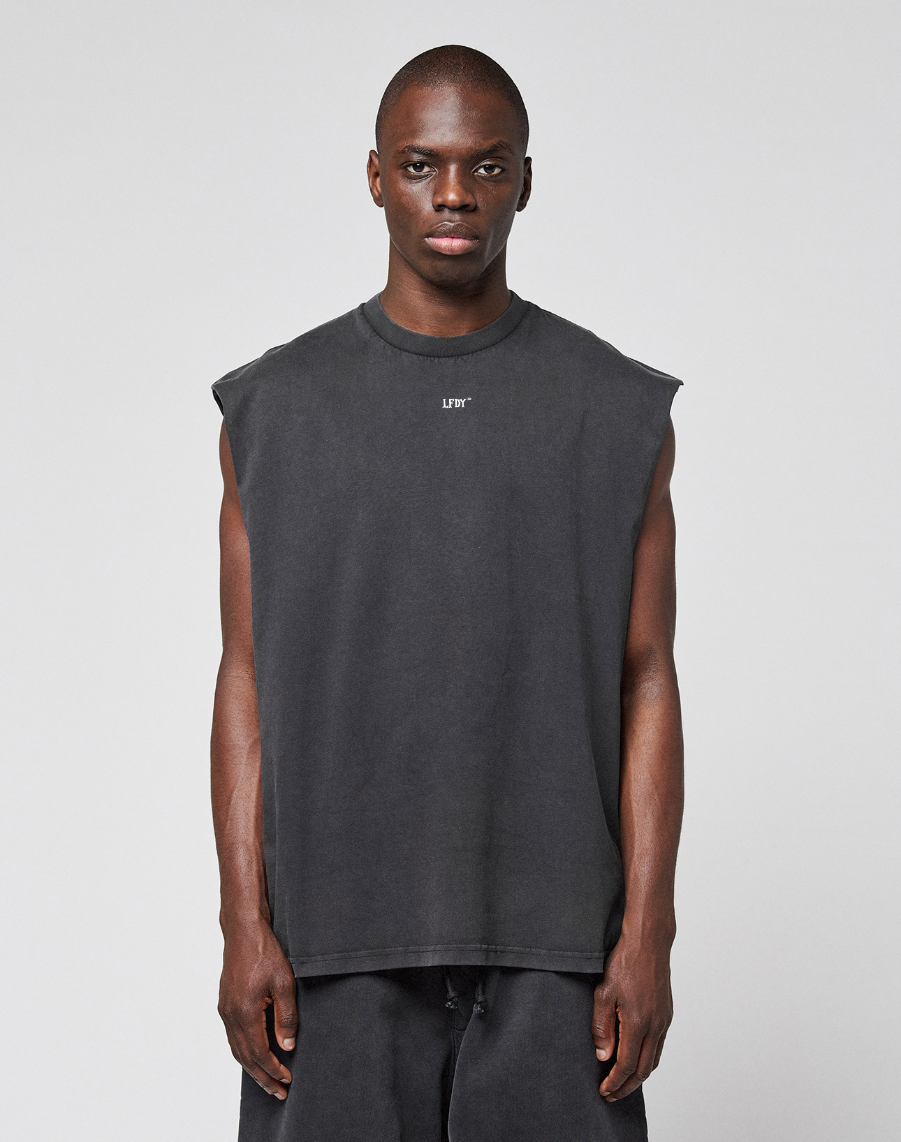 A person faces forward in an oversize fit LFDY Run Club Muscle Shirt by LIVE FAST DIE YOUNG, sleeveless and dark gray with matching pants, made from 100% Baumwolle and featuring a small white chest logo, against a plain light gray background.