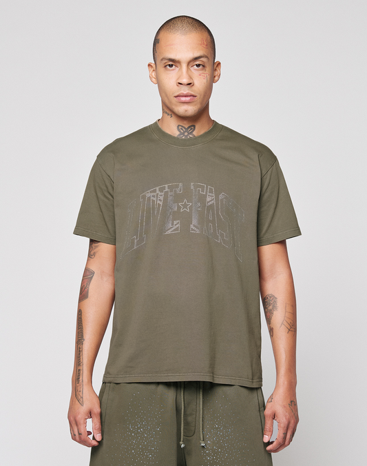 A tattooed person is wearing the LIVE FAST DIE YOUNG Live Fast Rhinestone Tee in olive green with matching shorts, standing against a plain light gray background. | model