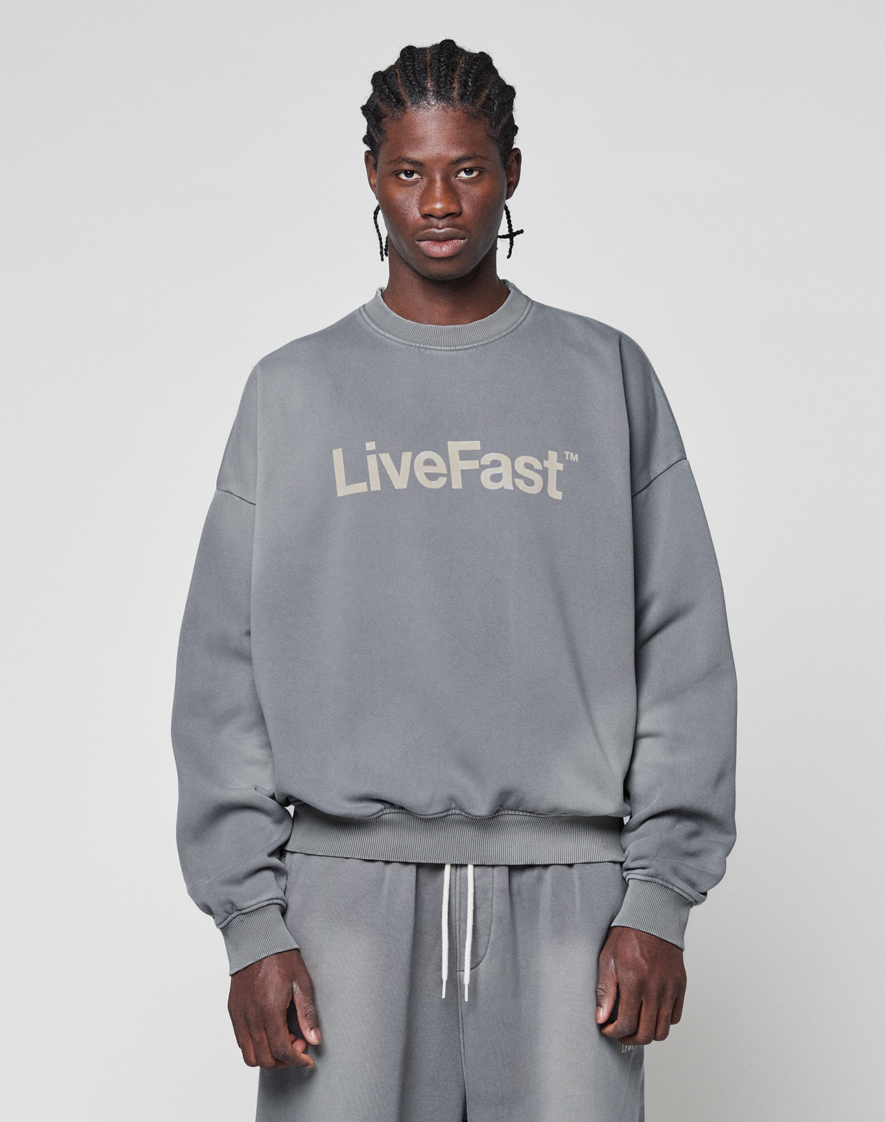 A person faces forward in a Basic LiveFast Sweater by LIVE FAST DIE YOUNG and matching gray sweatpants, both with a subtle faded look and the bold "LiveFast" logo, set against a plain light gray background.