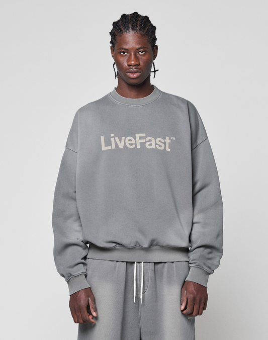 A man with braided hair wears the Basic LiveFast Sweater by LIVE FAST DIE YOUNG, an oversize fit gray sweatshirt with a handgefertigter Fade-Effekt and "LiveFast" print, paired with gray sweatpants against a light gray background. | model