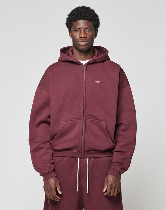 A man faces the camera in a maroon LIVE FAST DIE YOUNG Basic Hooded Zip with an oversized fit and matching sweatpants. Subtle branding details stand out against a plain, light gray background. | model