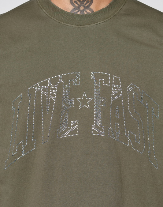 The Live Fast Rhinestone Tee by LIVE FAST DIE YOUNG features an olive green design with a large, silver rhinestone "LIVE FAST" logo across the chest. A tattoo on the bottom of a person's neck is visible at the top of the image.