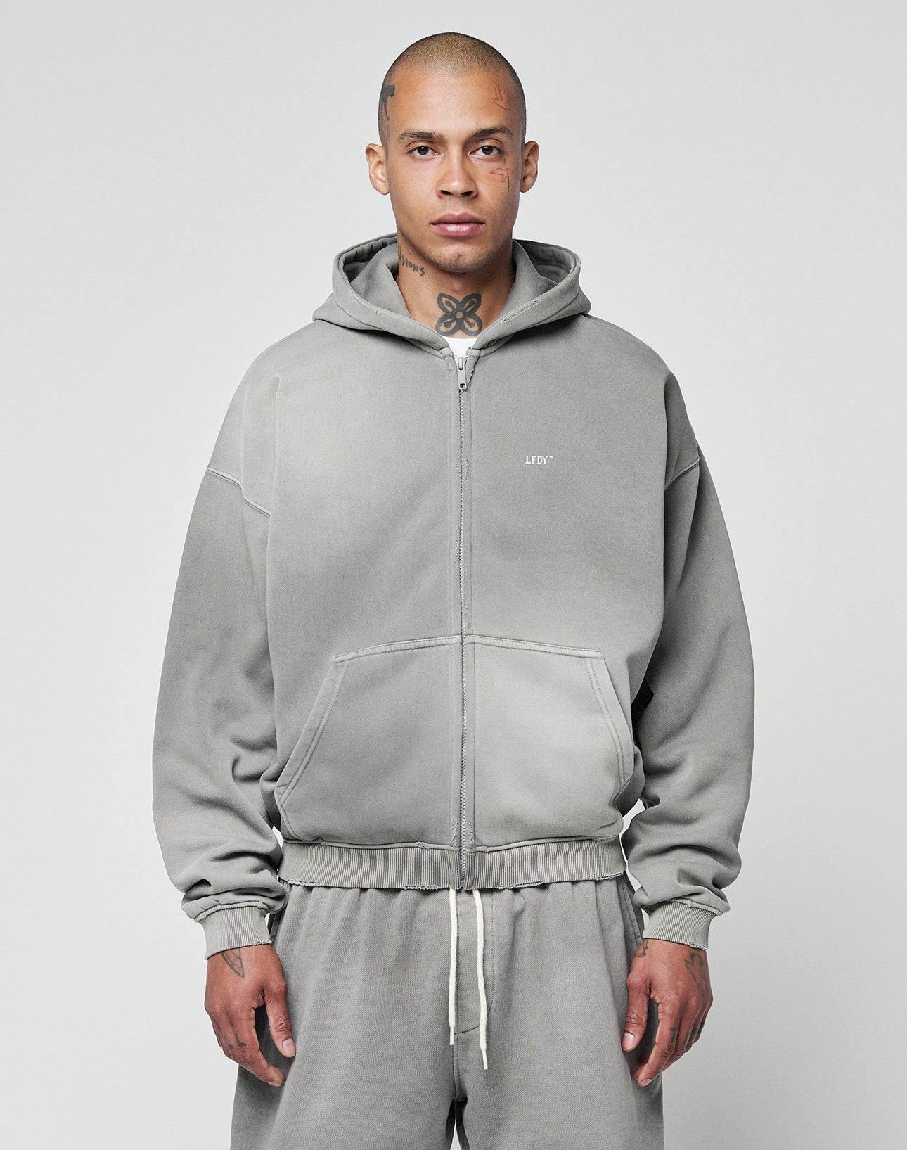 A person with a shaved head and neck tattoos models the LIVE FAST DIE YOUNG Basic Hooded Zip in gray, along with matching sweatpants, standing against a plain light background and looking at the camera.