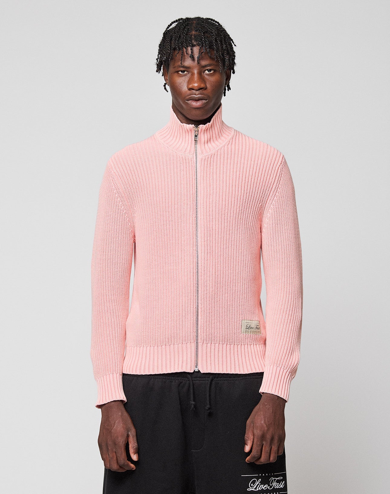 A man with short, textured hair wears the LIVE FAST DIE YOUNG LFDY Knit Jacket in light pink with ribbed texture and Stehkragen, paired with black pants, standing against a plain light gray background.