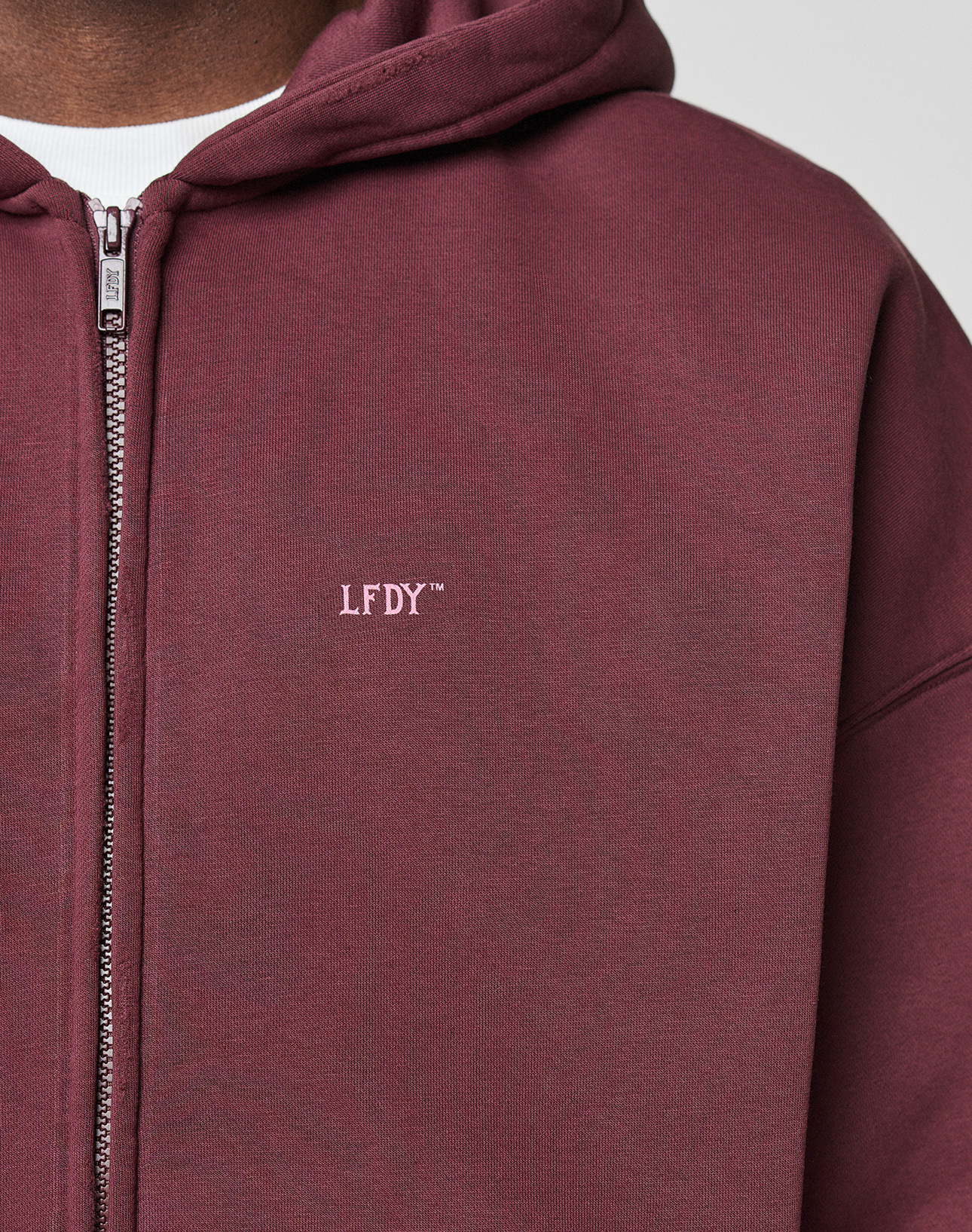 A person wears the LIVE FAST DIE YOUNG Basic Hooded Zip, a maroon zip-up hoodie with branding and logo on the chest, layered over a white shirt. The photo highlights the oversized fit, showing only their torso and part of their face.