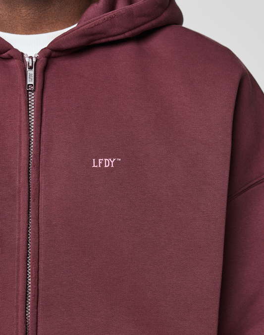 A person wears the LIVE FAST DIE YOUNG Basic Hooded Zip, a maroon zip-up hoodie with branding and logo on the chest, layered over a white shirt. The photo highlights the oversized fit, showing only their torso and part of their face.