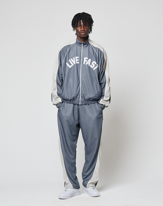 A person stands against a plain background wearing LIVE FAST DIE YOUNG’s gray and white LFDY DY Trackpants with a comfortable elastikbund and wide fit, paired with white sneakers.