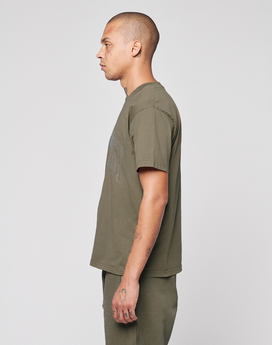 A person with short hair, wearing the LIVE FAST DIE YOUNG Live Fast Rhinestone Tee in olive-green oversize fit and matching pants, stands in profile facing left against a plain light background. Tattoos are visible on their arms and head.