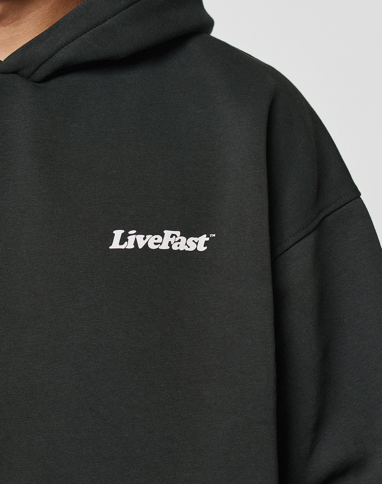 A close-up of the black Basic 360 Hooded by LIVE FAST DIE YOUNG in an oversize fit, featuring white LiveFast text on the left chest. The background is plain, showing part of the persons neck and chin.