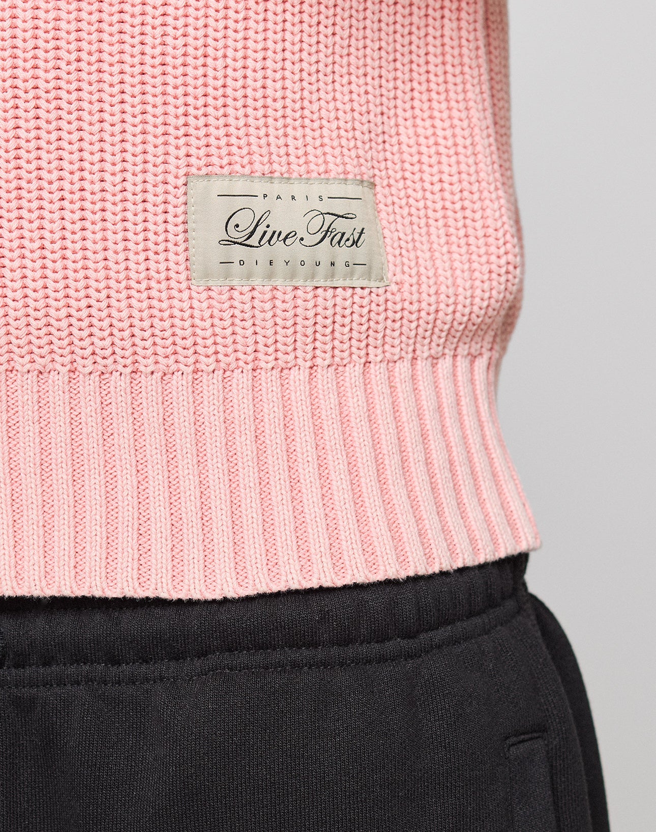 A close-up of the pink LFDY Knit Jacket by LIVE FAST DIE YOUNG, featuring a beige "Paris Live Fast Devour" label, 100% Baumwolle fabric, a Stehkragen, and styled with black pants.