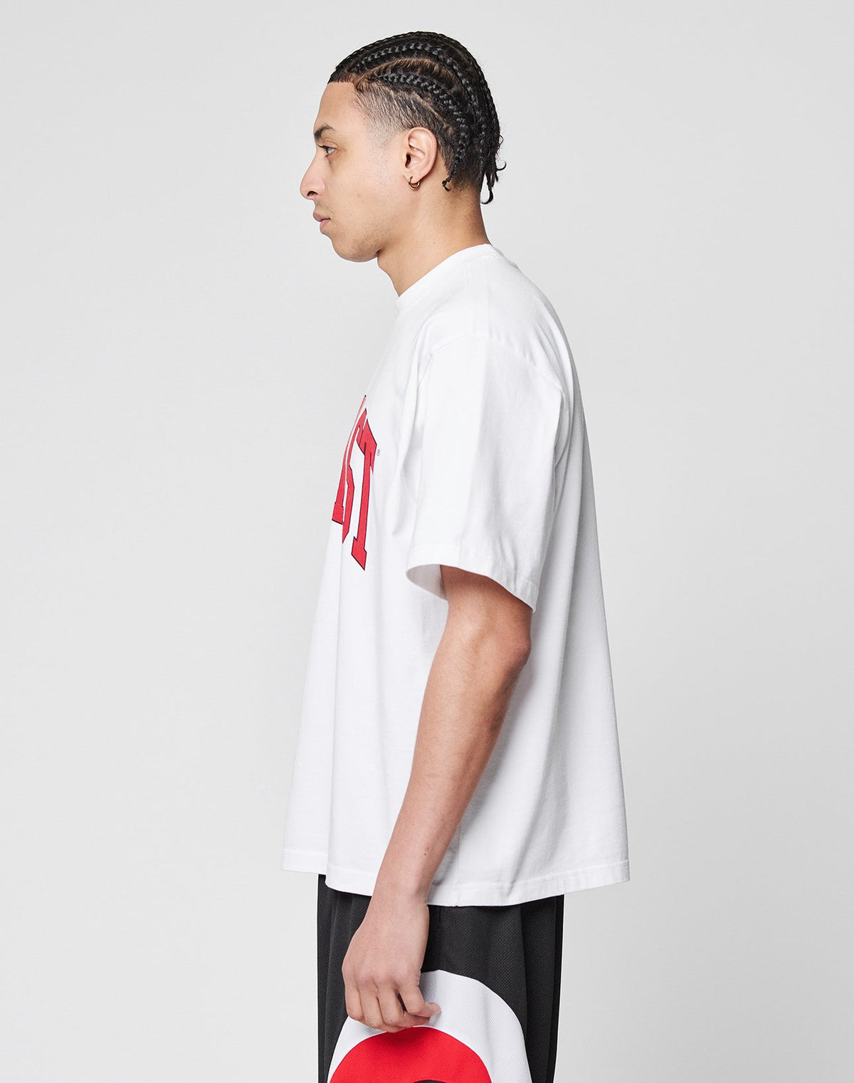 A person with braided hair stands in profile against a plain background, wearing the Live Fast College Tee—an oversize white T-shirt with red frontprint text—paired with black shorts and holding a white and red hat by their side.
