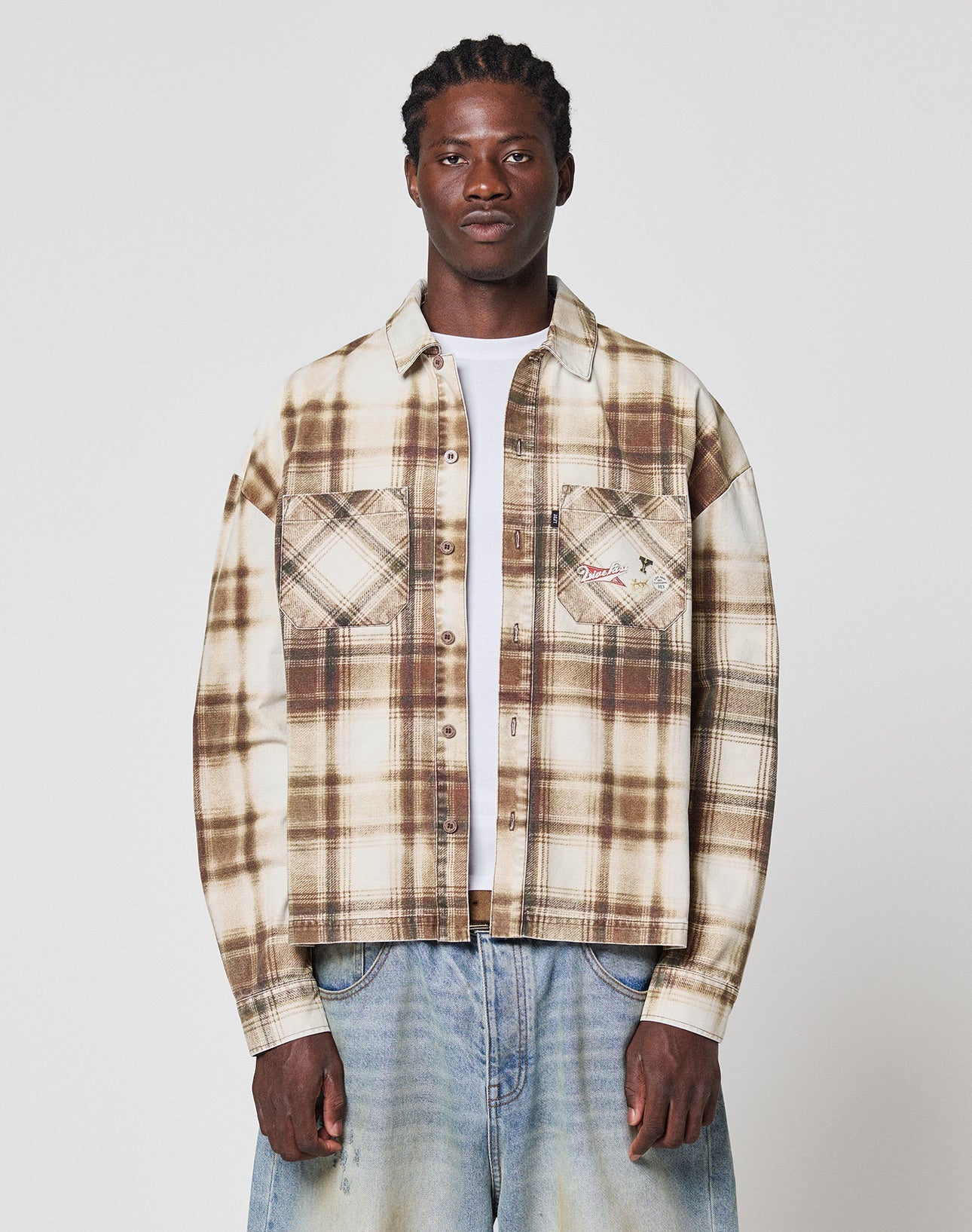 A person faces forward against a plain white background, wearing LIVE FAST DIE YOUNG’s Faded Flannel in beige and brown plaid with two chest pockets and oversize fit, paired with light blue baggy jeans—all made from soft Baumwolle.