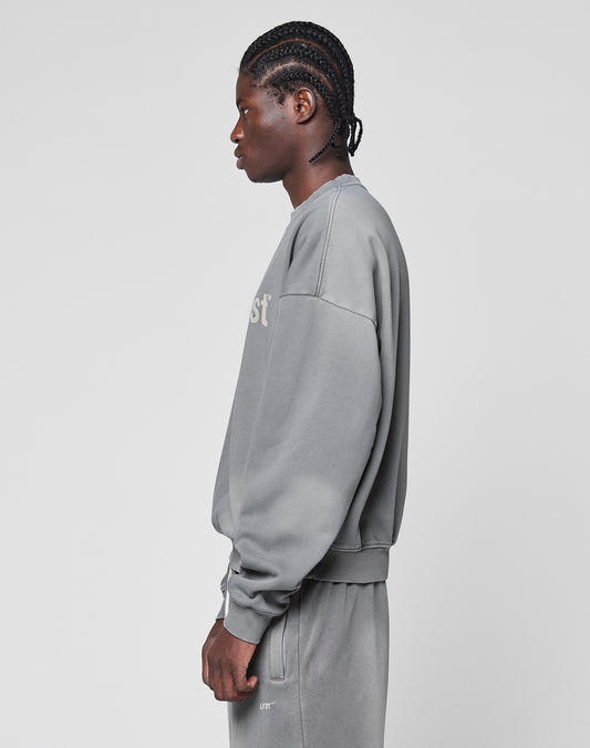 A person with braided hair stands sideways in an oversize, grey Basic LiveFast Sweater and matching sweatpants by LIVE FAST DIE YOUNG, featuring a subtle faded look against a plain white background.