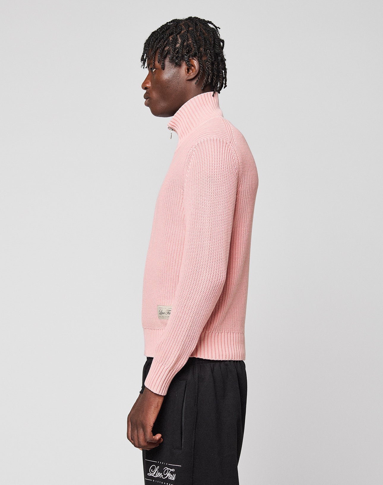 A person with short, textured hair wears a light pink LFDY Knit Jacket by LIVE FAST DIE YOUNG and black Regular Fit pants, standing in profile against a plain light gray background.