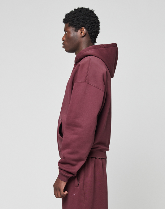 A person stands in profile against a plain background, wearing an oversize fit maroon LIVE FAST DIE YOUNG Basic Hooded Zip and matching sweatpants with subtle branding. The hood is down, hands at their sides.
