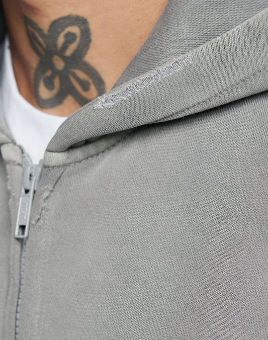 Close-up of a person wearing the LIVE FAST DIE YOUNG Basic Hooded Zip in grey, showing a slight tear on the hood edge and a subtle faded look. A white t-shirt underneath and part of a black neck tattoo are also visible.