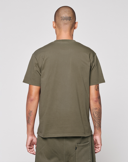 A person with short hair and visible tattoos stands facing away, wearing the LIVE FAST DIE YOUNG Live Fast Rhinestone Tee in olive green and matching shorts against a plain light background.