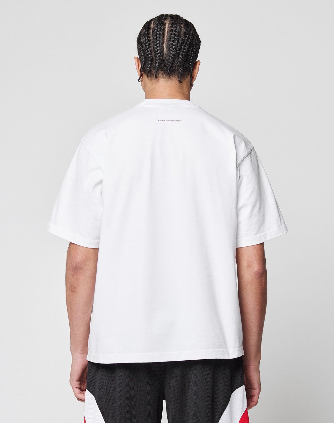 A person with braided hair stands with their back to the camera, wearing the Live Fast College Tee in plain white (100% Baumwolle) and black shorts with red and white patterns against a plain light gray background.