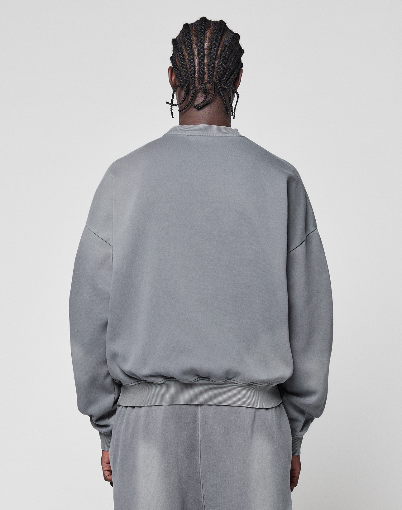 A person with cornrow braids and silver hoops wears the LIVE FAST DIE YOUNG Basic LiveFast Sweater in gray with matching faded-look pants, standing with their back to the camera against a plain light background.