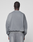 A person with cornrow braids and silver hoops wears the LIVE FAST DIE YOUNG Basic LiveFast Sweater in gray with matching faded-look pants, standing with their back to the camera against a plain light background.
