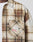 A close-up shows a person wearing the LIVE FAST DIE YOUNG Faded Flannel—a brown and beige plaid, oversize cotton shirt—over a white T-shirt. The shirt’s pocket is decorated with pins and patches; only the torso and part of the arm are visible.