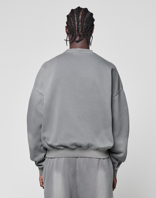 A person with braided hair stands with their back to the camera, wearing an oversize fit Basic LiveFast Sweater and matching gray pants by LIVE FAST DIE YOUNG, both in a subtle faded gray, set against a plain light background.