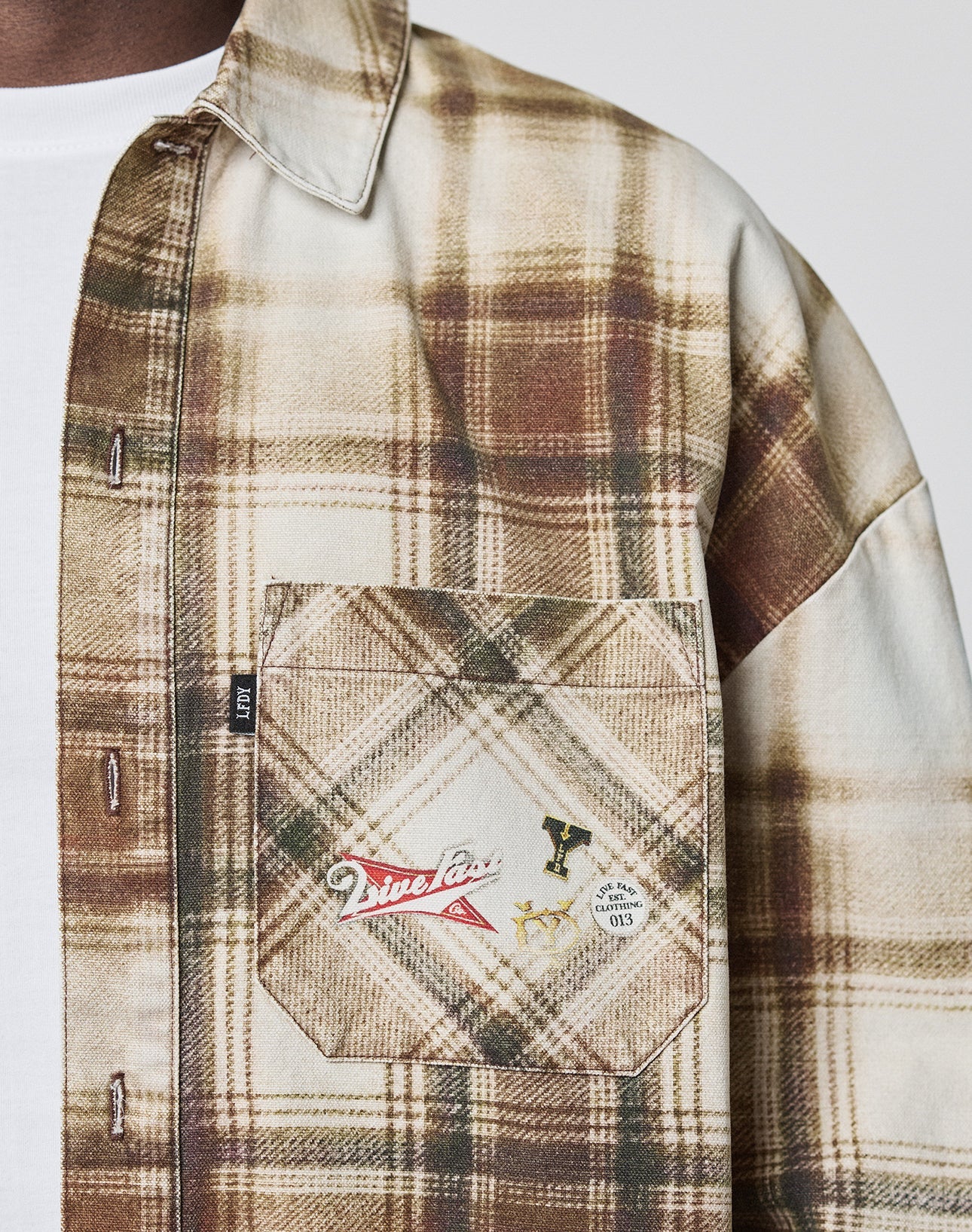 A close-up shows a person wearing the LIVE FAST DIE YOUNG Faded Flannel—a brown and beige plaid, oversize cotton shirt—over a white T-shirt. The shirt’s pocket is decorated with pins and patches; only the torso and part of the arm are visible.