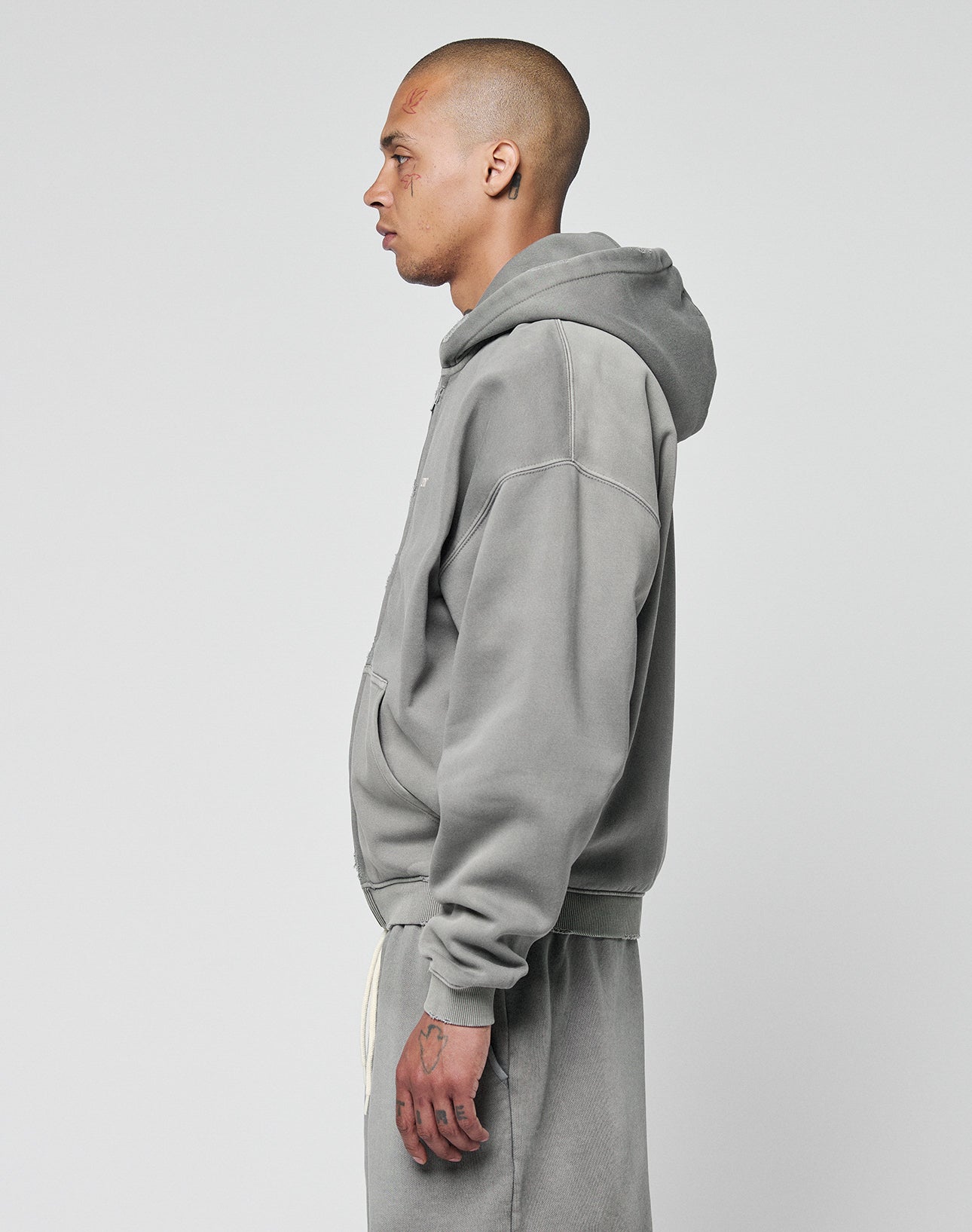 A person with a shaved head stands in profile, wearing the LIVE FAST DIE YOUNG Basic Hooded Zip hoodie and matching sweatpants with a subtle logo print and faded look, set against a plain light background.