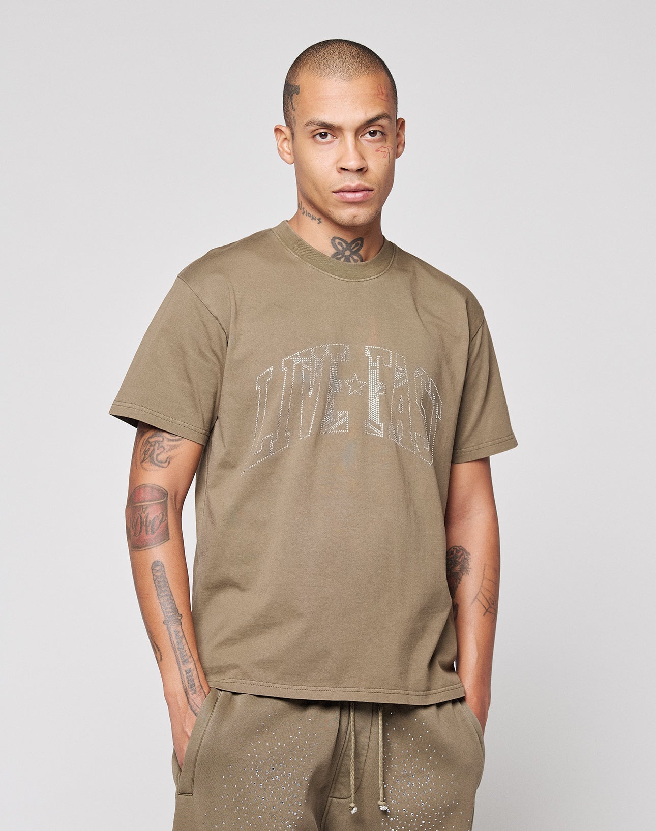 A tattooed man wears the LIVE FAST DIE YOUNG Live Fast Rhinestone Tee in an oversized khaki fit, paired with matching khaki pants. He stands facing forward with hands in pockets against a plain light background.