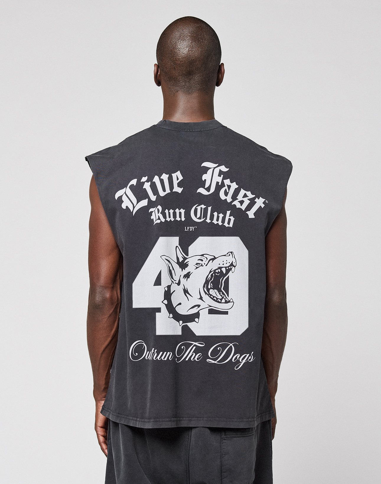 A person faces away, wearing the LFDY Run Club Muscle Shirt by LIVE FAST DIE YOUNG—an oversized, sleeveless black top with bold white "Live Fast Run Club" text, a large number 40, dog graphic, and "Outrun The Dogs" on the back for a vintage look. | model