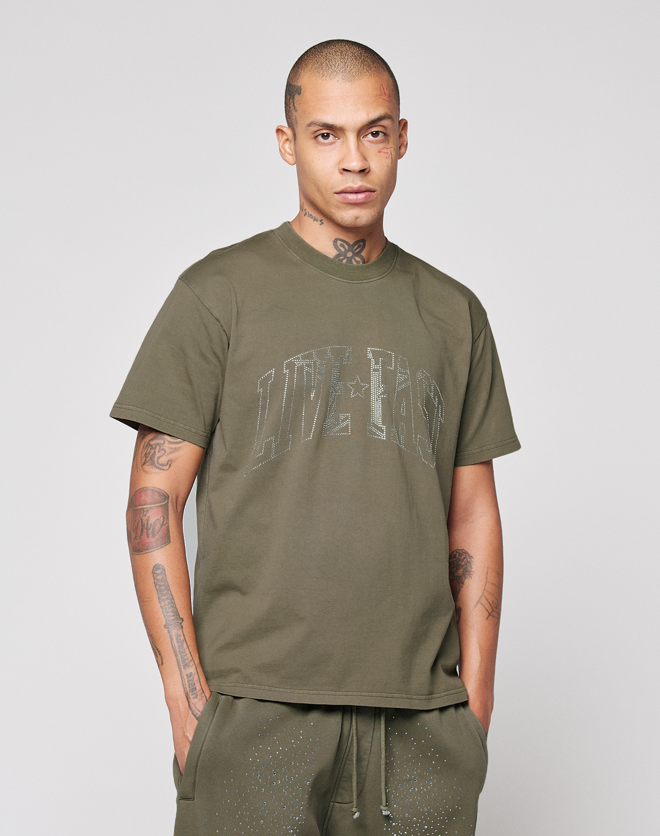 A man with neck and arm tattoos wears an oversized olive green LIVE FAST DIE YOUNG Live Fast Rhinestone Tee and matching pants, standing against a plain light gray background with a neutral expression and hands in his pockets.