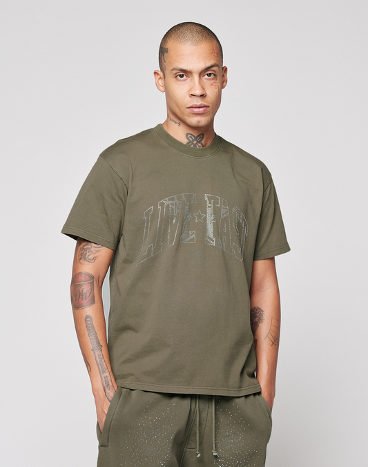 A man with neck and arm tattoos wears an oversized olive green LIVE FAST DIE YOUNG Live Fast Rhinestone Tee and matching pants, standing against a plain light gray background with a neutral expression and hands in his pockets.