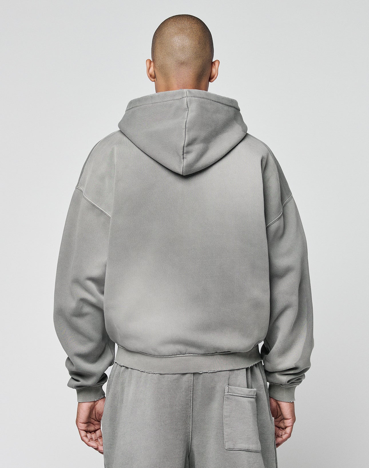 A person with a shaved head, dressed in the LIVE FAST DIE YOUNG Basic Hooded Zip and matching sweatpants with a subtle faded look, stands facing away from the camera against a plain light background.