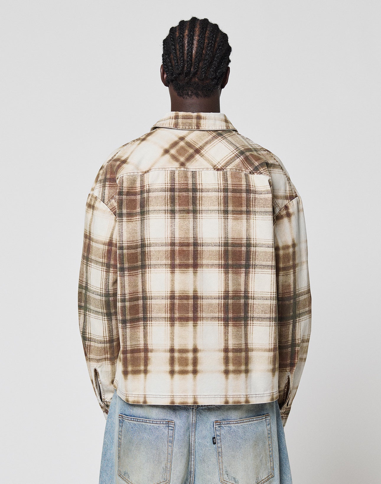 A person with braided hair, seen from behind, wears the LIVE FAST DIE YOUNG Faded Flannel in brown and cream plaid with light blue jeans, standing against a plain white background.