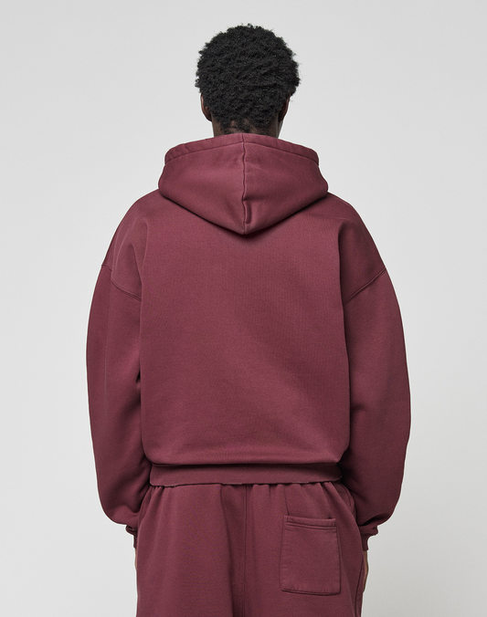 A person with short curly hair faces away, wearing a maroon LIVE FAST DIE YOUNG Basic Hooded Zip and matching sweatpants in an oversize fit, against a plain light gray background.