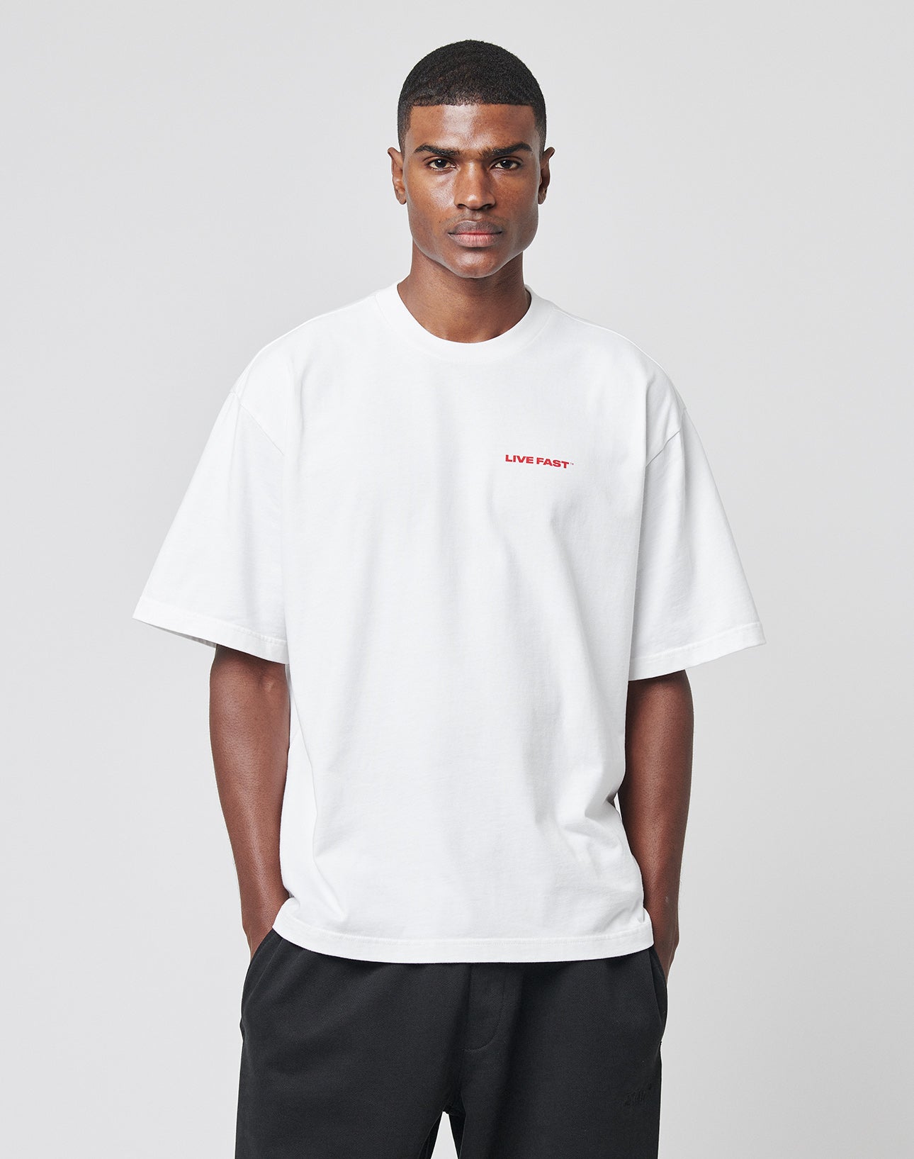A man stands against a plain light gray background with his hands in his pockets, wearing the LIVE FAST DIE YOUNG LFDY Cinema Tee—an oversize fit 100% cotton white t-shirt with LIVE FAST. printed in small red letters on the left chest.