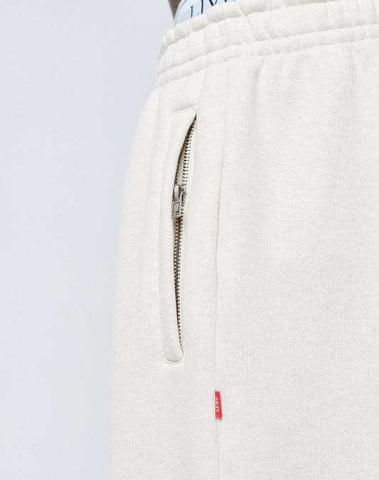 Close-up of cream-colored LFDY Heavyweight Loungepants by LIVE FAST DIE YOUNG, made from 650gsm cotton with an elastic waistband, metal zippered pocket, red fabric tag at the seam, and a white waistband with black text visible at the top.