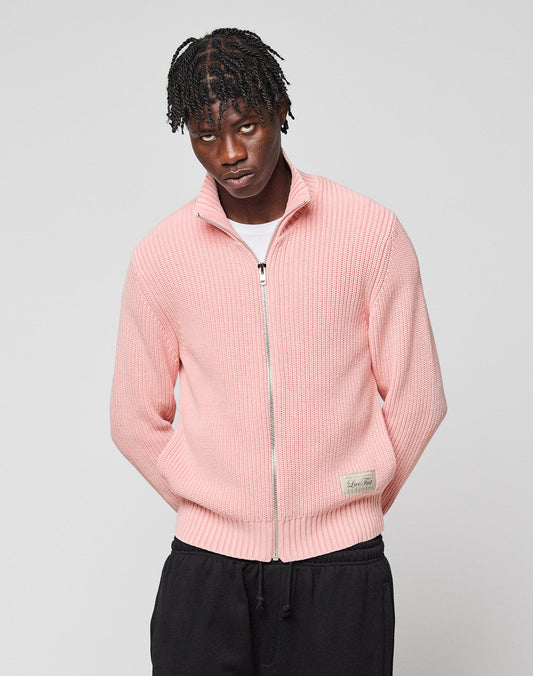 A man with braided hair wears the LIVE FAST DIE YOUNG LFDY Knit Jacket in light pink over a white t-shirt and black pants, standing against a plain light gray background.
