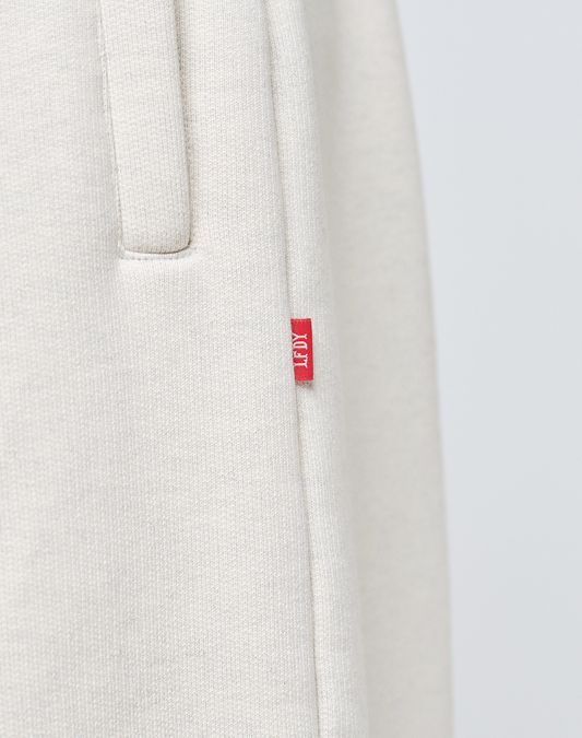 Close-up of LFDY Heavyweight Loungepants by LIVE FAST DIE YOUNG in light beige, featuring textured Baumwollegewebe, a pocket, and red AIM tag. Crafted from 650gsm heavyweight fabric for a premium feel inspired by classic Wide Fit Jogger styles.
