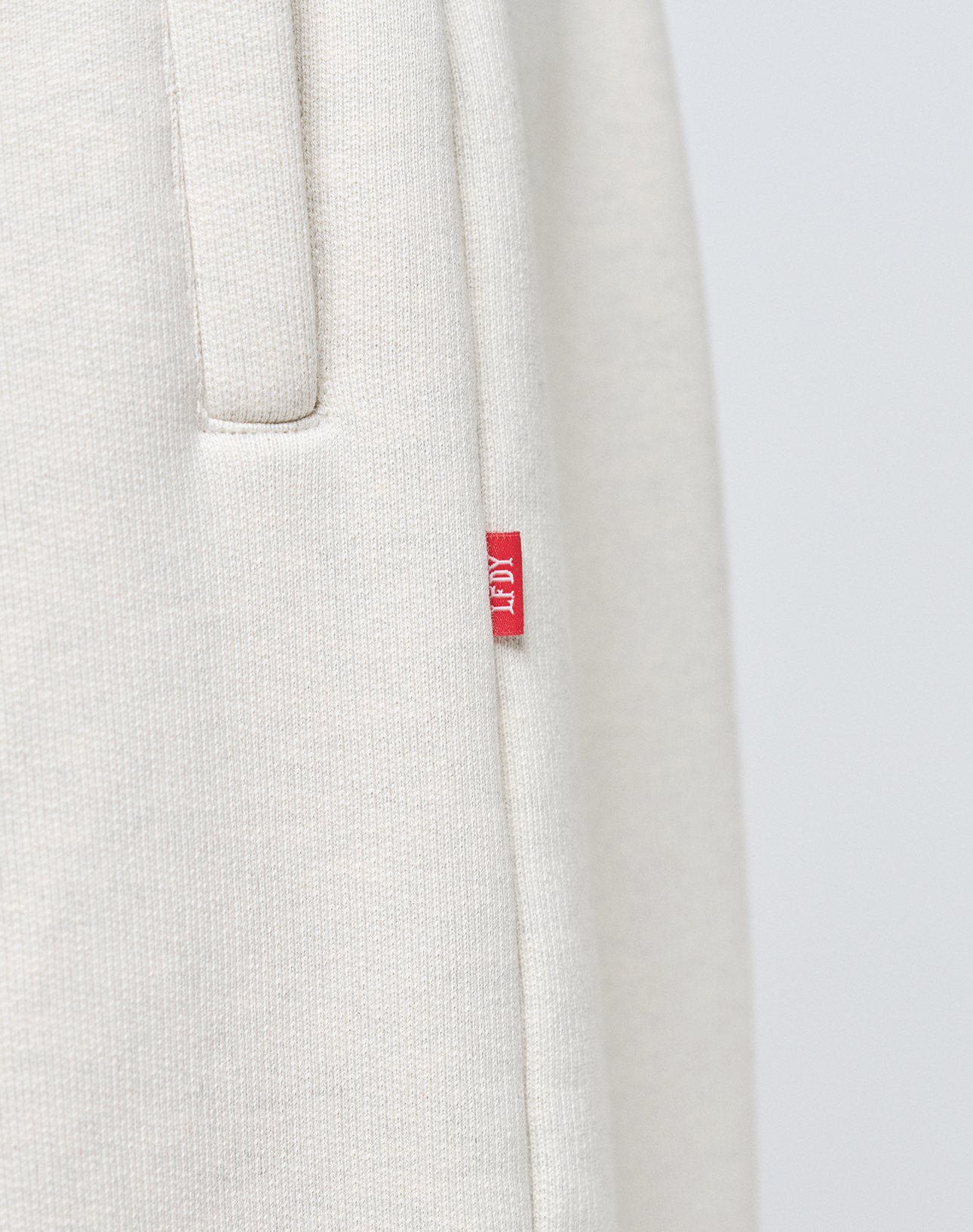 Close-up of LFDY Heavyweight Loungepants by LIVE FAST DIE YOUNG in light beige, featuring textured Baumwollegewebe, a pocket, and red AIM tag. Crafted from 650gsm heavyweight fabric for a premium feel inspired by classic Wide Fit Jogger styles.
